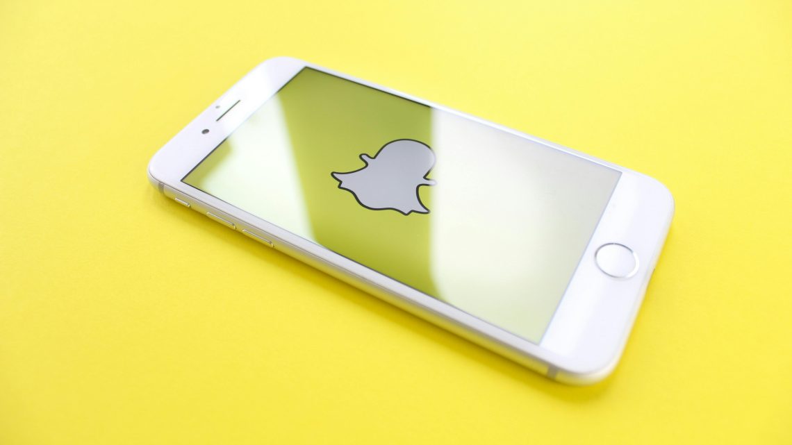 White smartphone displaying Snapchat logo on a bright yellow background, angled view.
